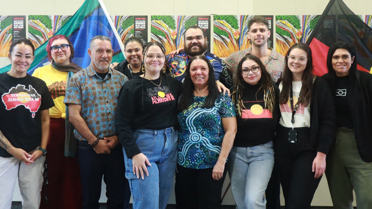 A group of 11 men and women stand in front of the Aboriginal and Torres Strait Islander flags. Posters from NAIDOC Week 2024 run in a line on the wall behind them