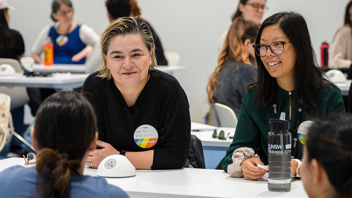 Two female staff members sit at a table in a room full of people