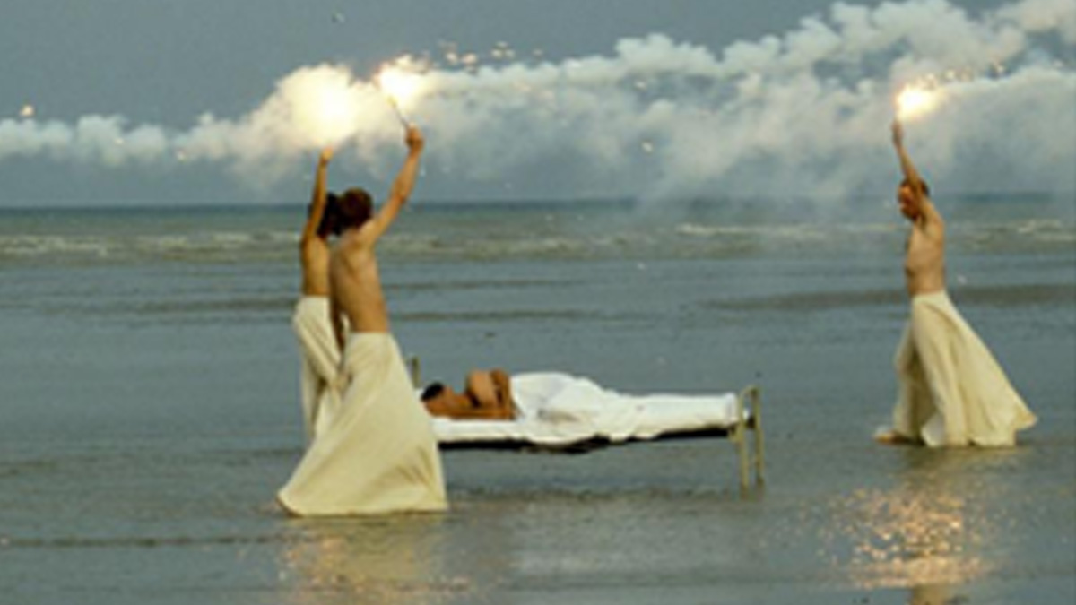 Shirtless people holding flares walking around a bed at the sea's edge