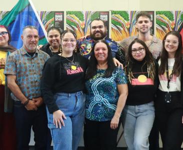 A group of 11 men and women stand in front of the Aboriginal and Torres Strait Islander flags. Posters from NAIDOC Week 2024 run in a line on the wall behind them