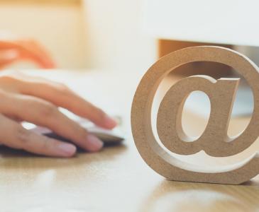 A wooden @ symbol on a table with a person's hands using a mouse in the background